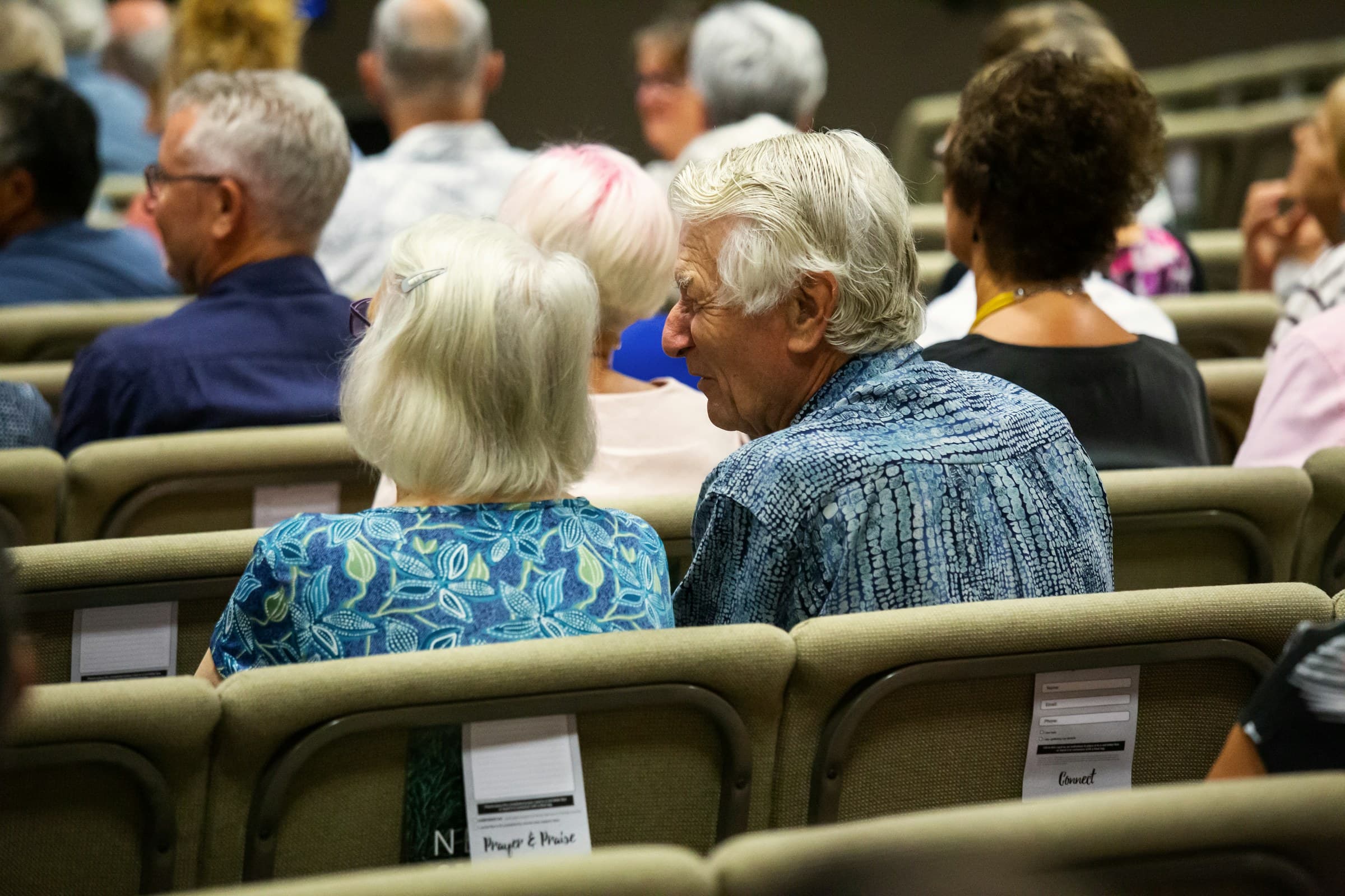 Ältere Menschen sitzen als Publikum in einem Saal und unterhalten sich